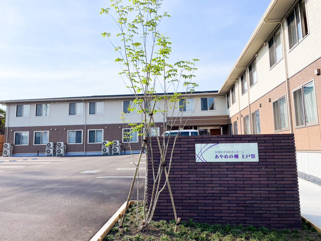 介護施設の外観。白と茶色の2階建て建物、駐車場、新しい植栽が見える